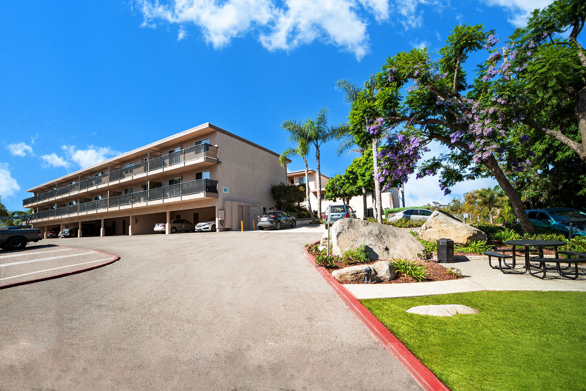 Three-story apartment complex with covered ground-level parking under a bright blue sky.